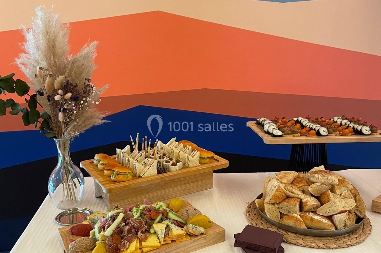 Table dressée avec des plateaux de fromages, charcuteries, sushis et pains, devant un mur aux motifs colorés.