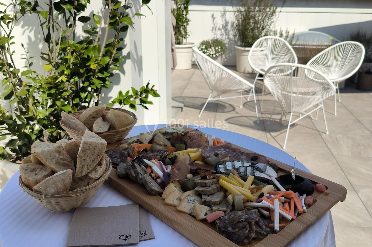 Plateau de charcuterie et fromages avec légumes et pain, posé sur une table dressée en extérieur, près de chaises et plantes.