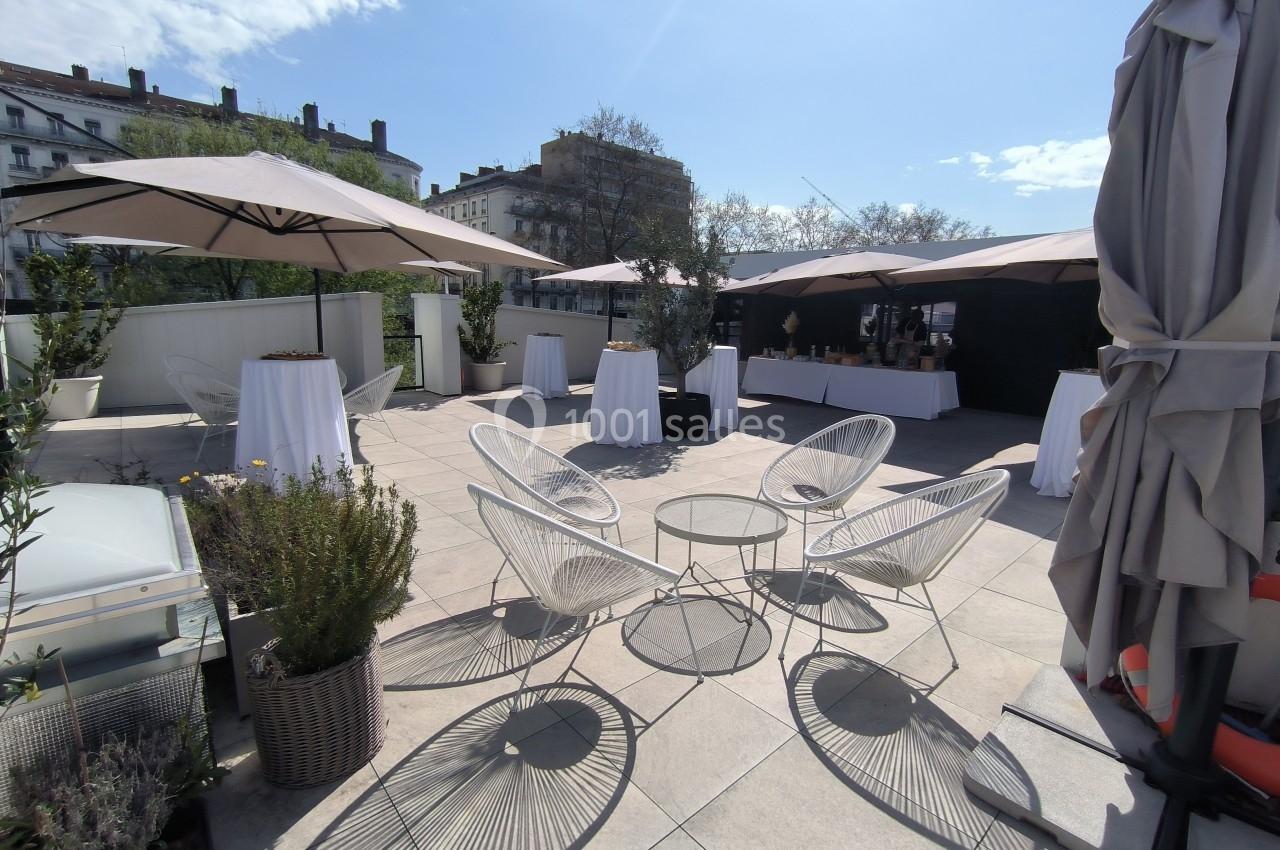 Terrasse aménagée avec des tables hautes, des chaises design, des parasols et des plantes sous un ciel ensoleillé.