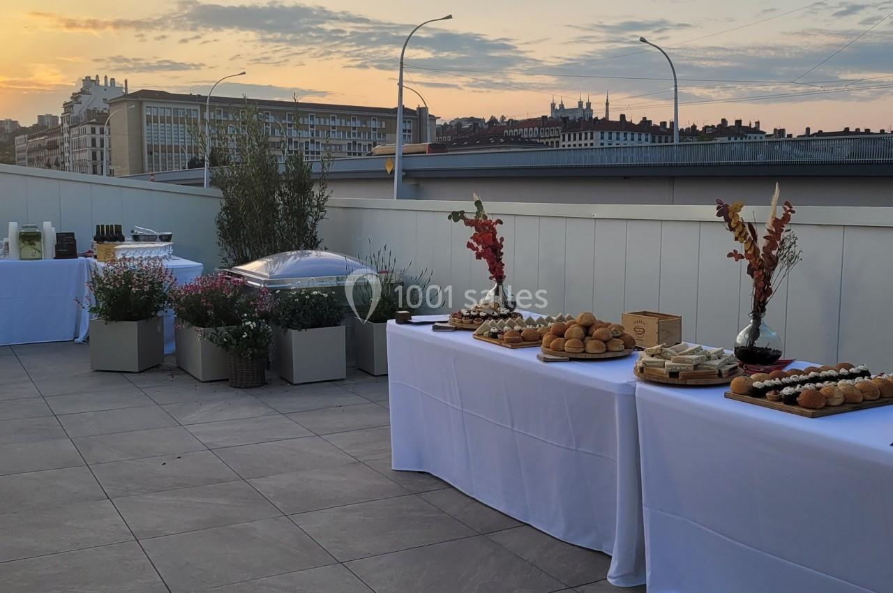 Buffet dressé sur une terrasse au coucher du soleil, avec vue sur des bâtiments urbains en arrière-plan.