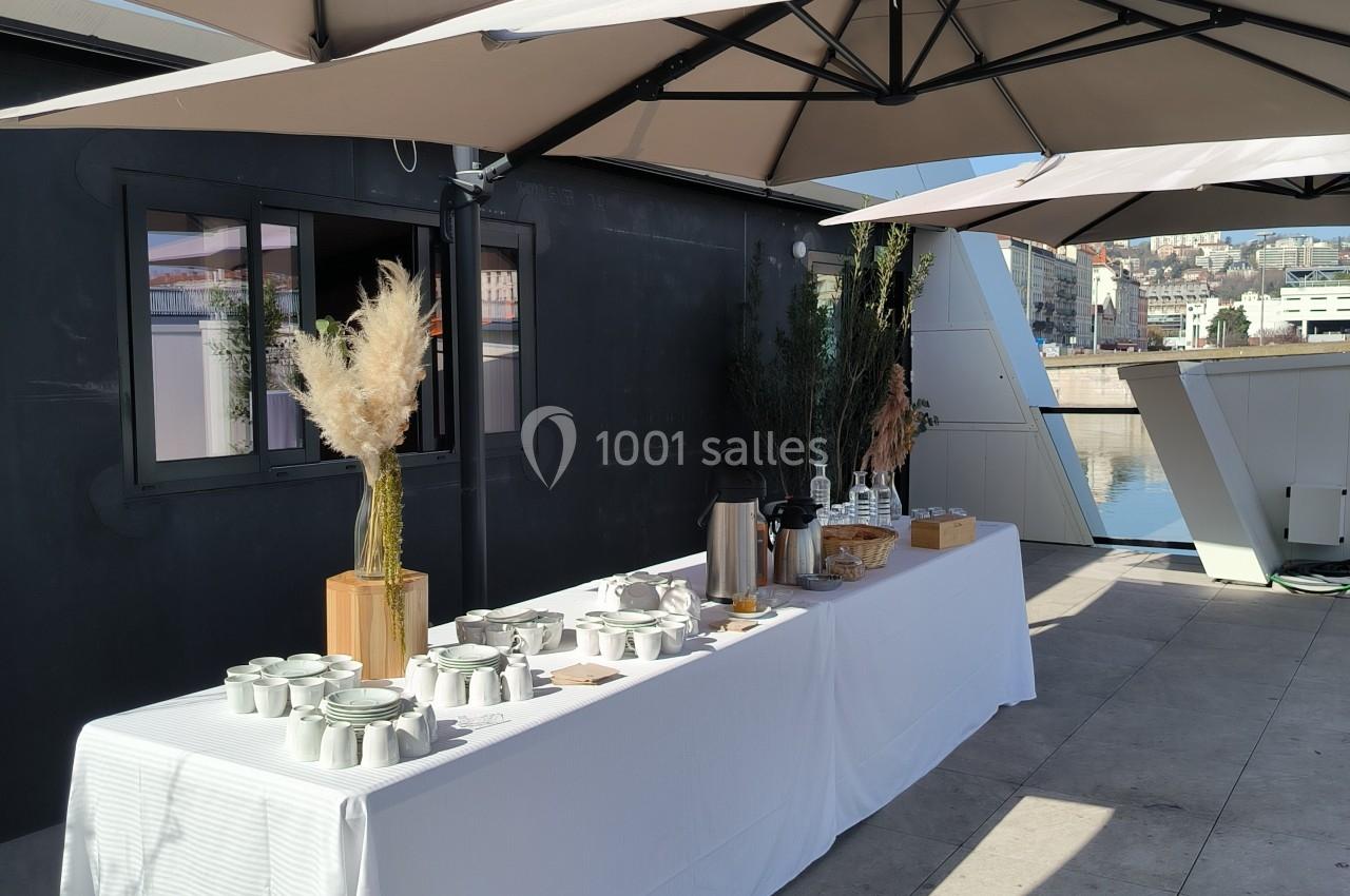 Table dressée en extérieur sous des parasols, avec vaisselle, thermos et décorations végétales.