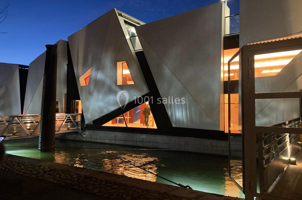 Bâtiment moderne aux façades angulaires éclairées, situé au bord d'un canal, photographié à la tombée de la nuit.