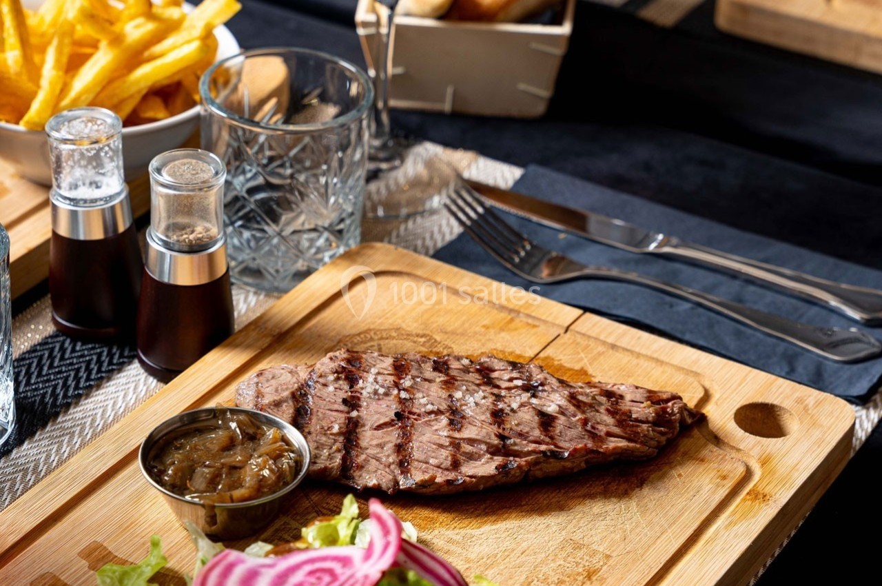 Steak grillé servi sur une planche en bois avec salade, oignons confits et frites en arrière-plan.