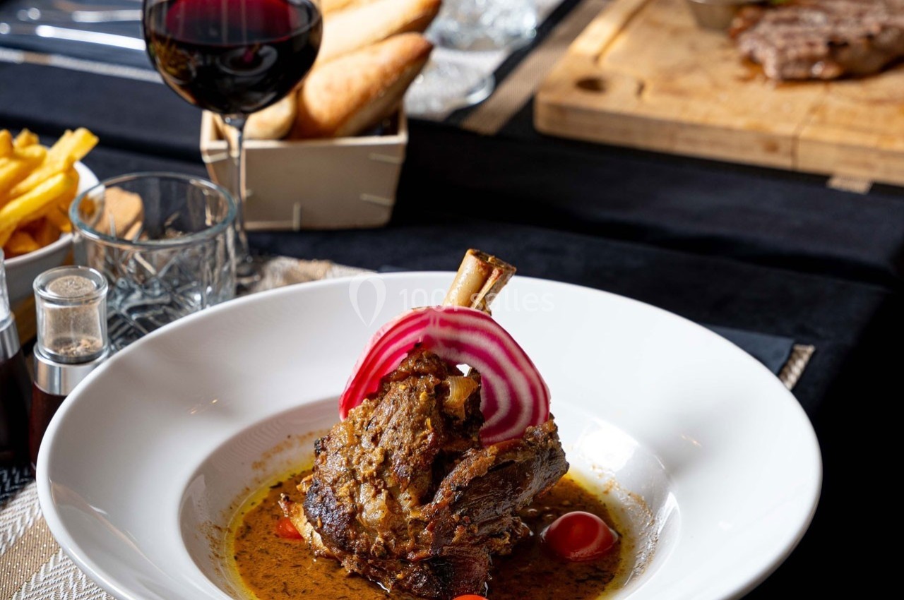 Plat de jarret d'agneau braisé servi avec sauce, rondelles d'oignon rouge et tomates cerises, accompagné de vin rouge.