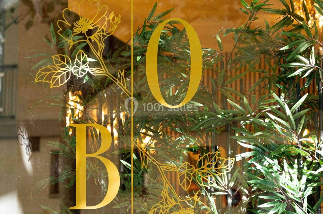 Lettrage doré ’B’ et ’O’ sur une vitre, entouré de motifs floraux et de plantes vertes en arrière-plan.