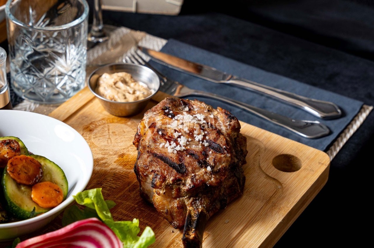Côte de porc grillée avec sel, accompagnée de légumes, sauce et couverts sur une planche en bois.