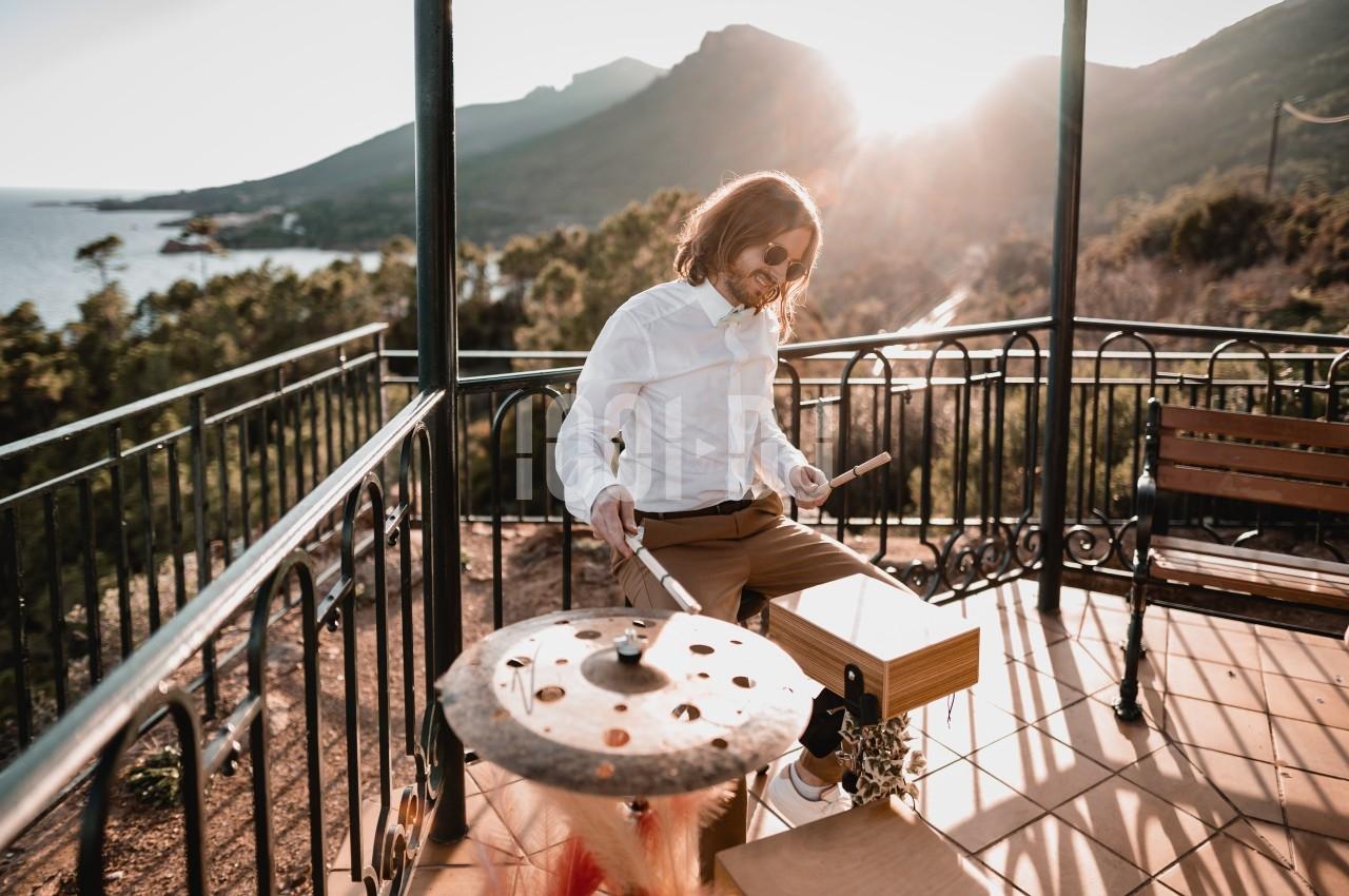 Un homme joue de la batterie sur une terrasse ensoleillée avec vue sur des montagnes et la mer.