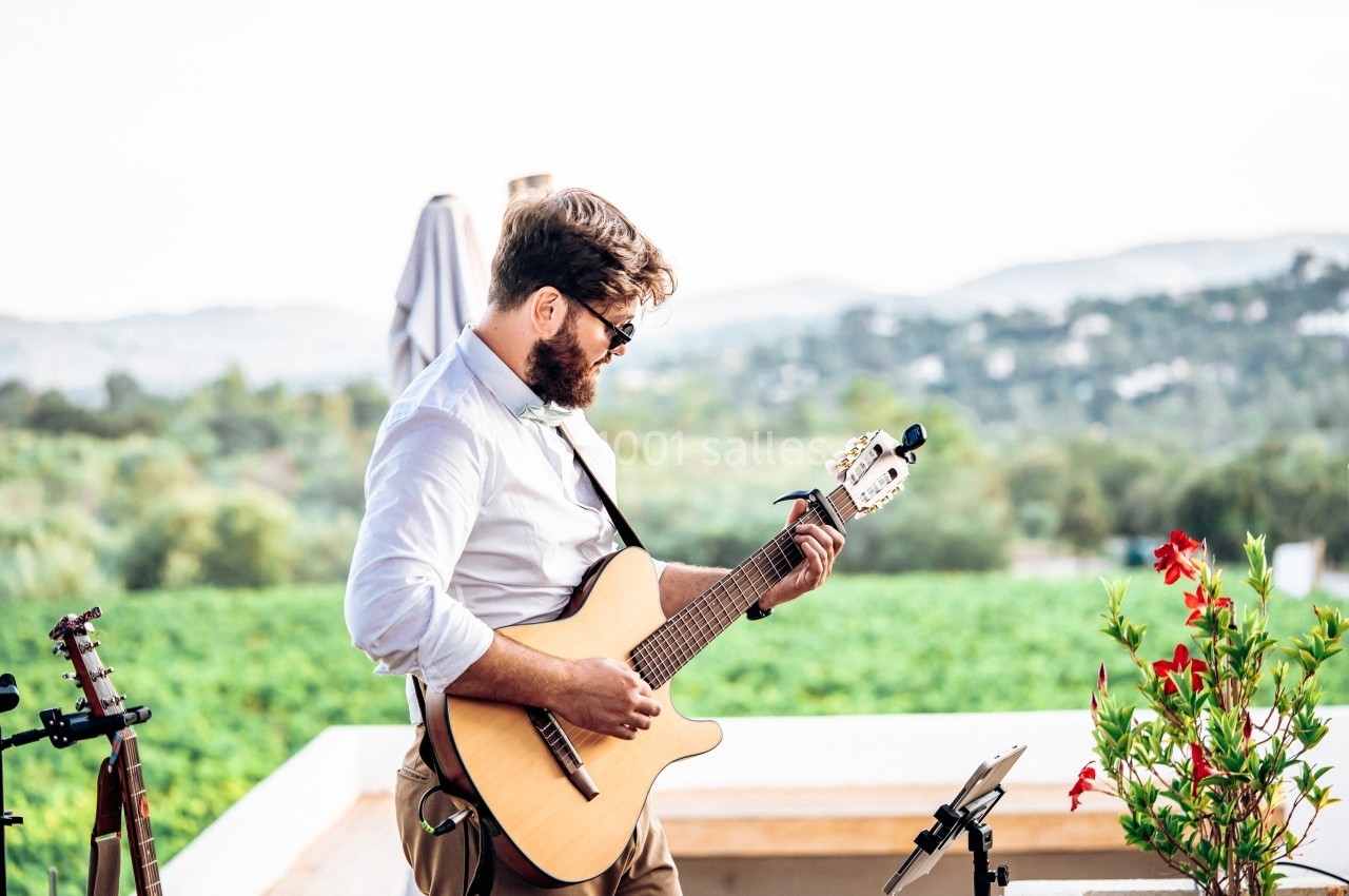 Un homme joue de la guitare acoustique en extérieur, avec un paysage verdoyant en arrière-plan.