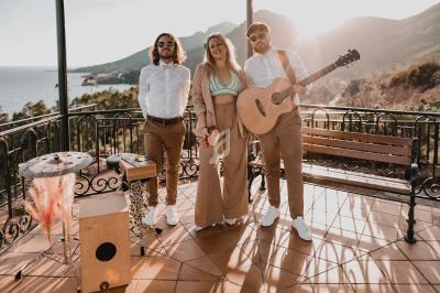Trois musiciens posent sur une terrasse ensoleillée avec vue sur la mer et les montagnes, guitare et percussions visibles.