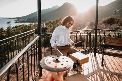 Un homme joue de la batterie sur une terrasse ensoleillée avec vue sur des montagnes et la mer.