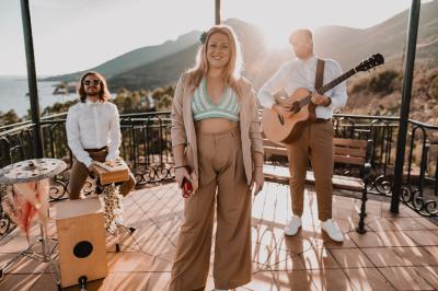 Une chanteuse souriante entourée de deux musiciens jouant de la guitare et du cajón sur une terrasse ensoleillée.