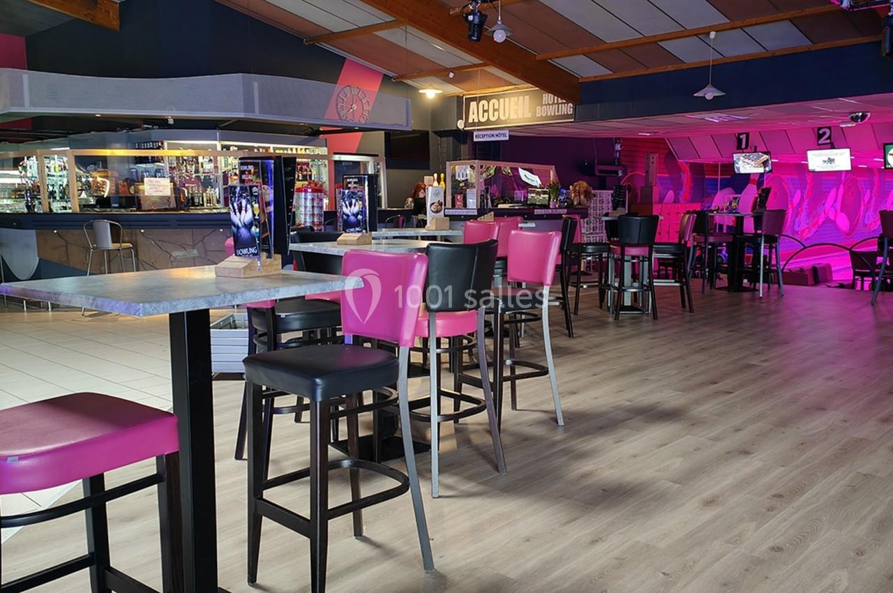 Salle de loisirs avec tables, chaises roses et noires, comptoir d'accueil et écrans lumineux en arrière-plan.