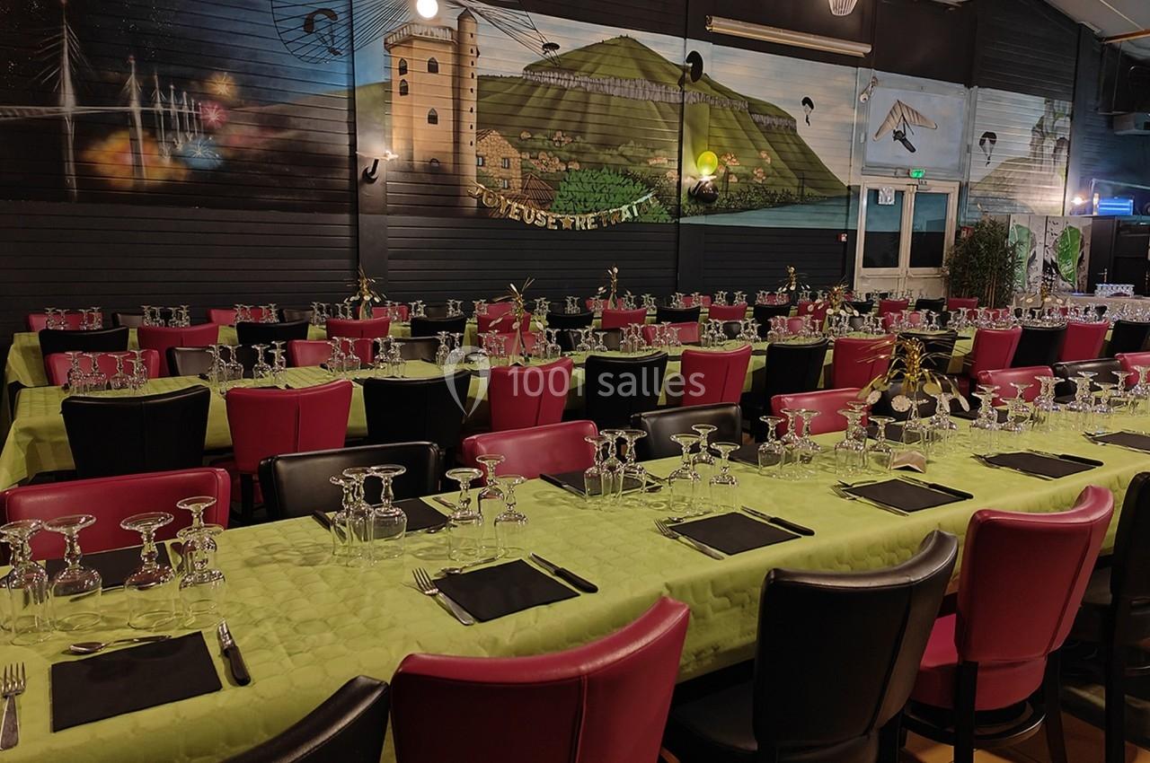 Salle de réception avec de longues tables dressées, nappes vertes, chaises colorées et fresque murale en arrière-plan.