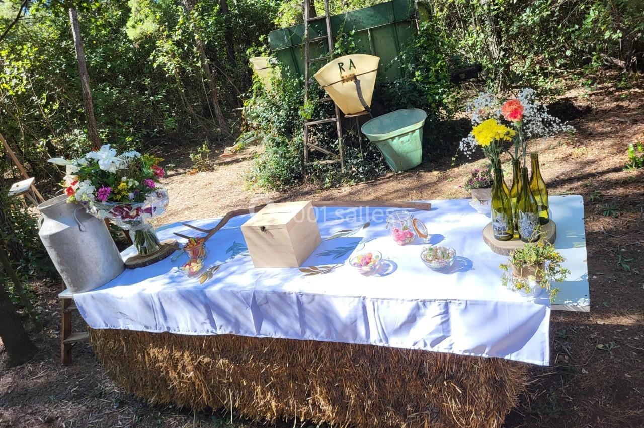 Table décorée avec des fleurs, des bouteilles et des objets rustiques, posée sur une botte de paille dans un cadre boisé.