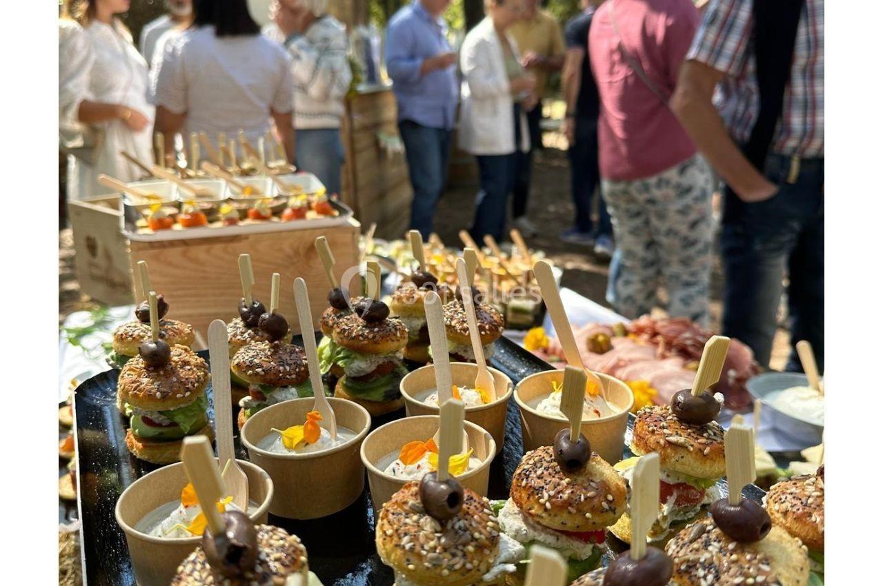 Plateau de mini-burgers et verrines garnies, présentés lors d'un buffet en extérieur avec des convives en arrière-plan.