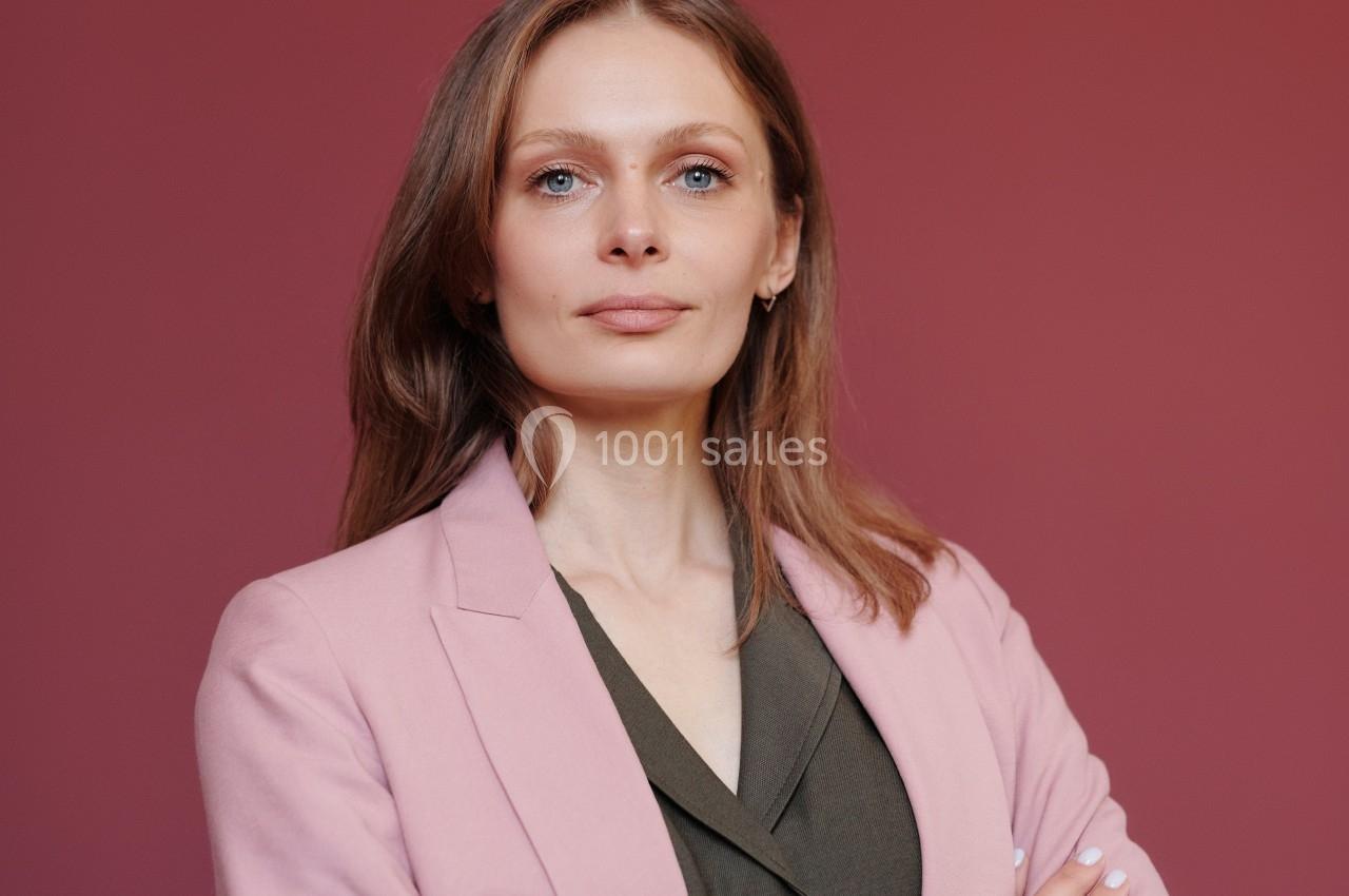 Femme aux cheveux châtains portant un blazer rose et une chemise kaki, posant devant un fond rose uni.