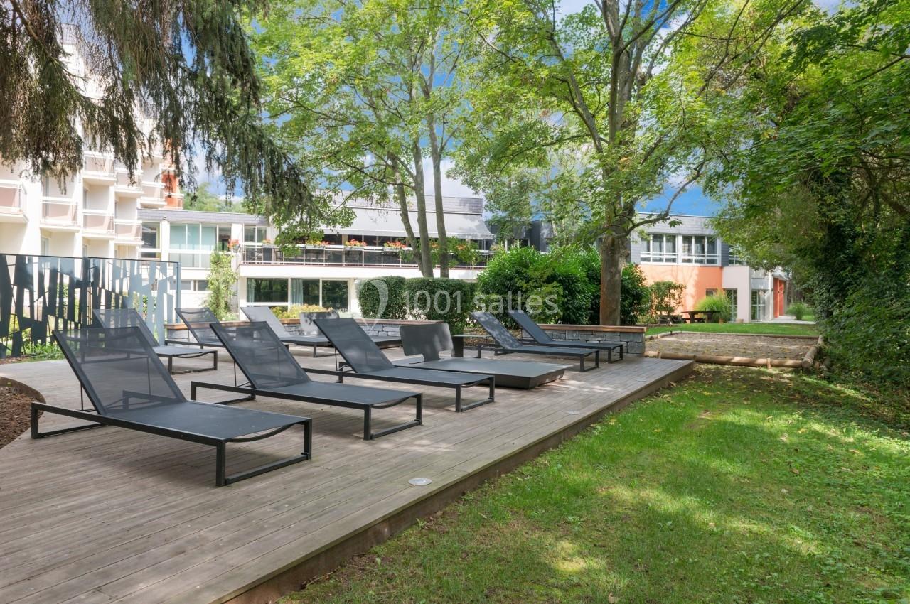 Chaises longues noires disposées sur une terrasse en bois entourée d'arbres et de bâtiments modernes.