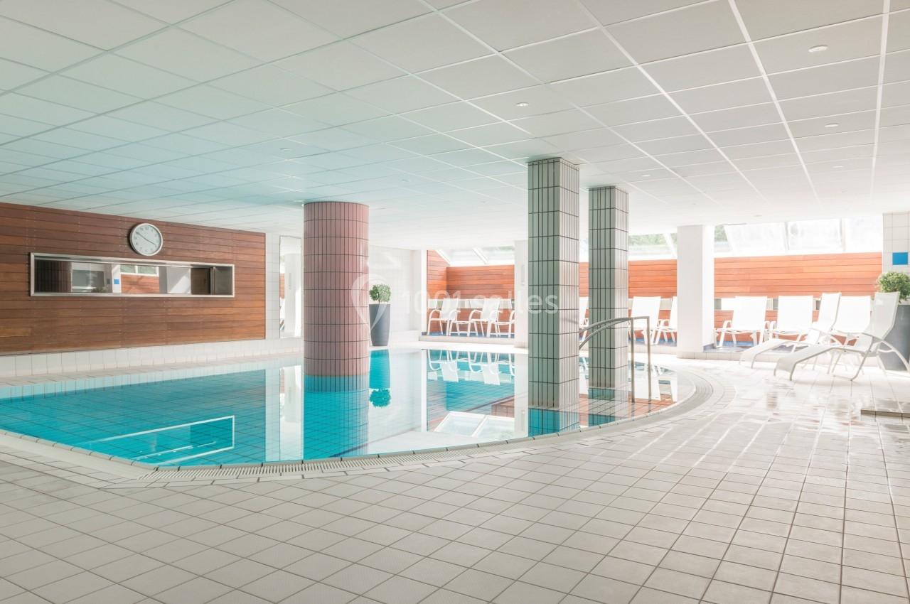 Piscine intérieure lumineuse entourée de chaises longues, avec murs en bois et carrelage blanc.