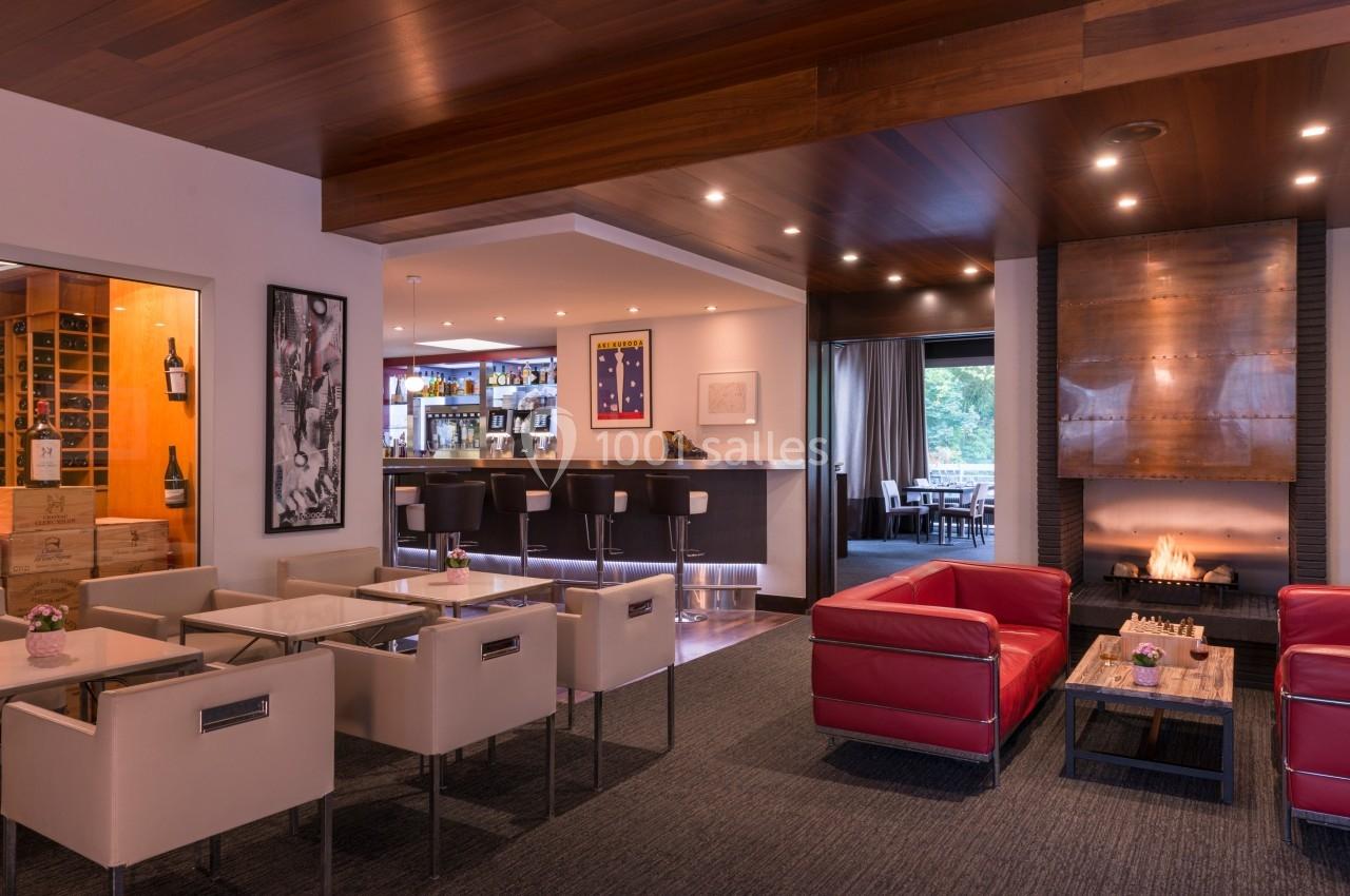 Salon moderne avec fauteuils rouges et blancs, cheminée, bar en arrière-plan et grande baie vitrée donnant sur l'extérieur.