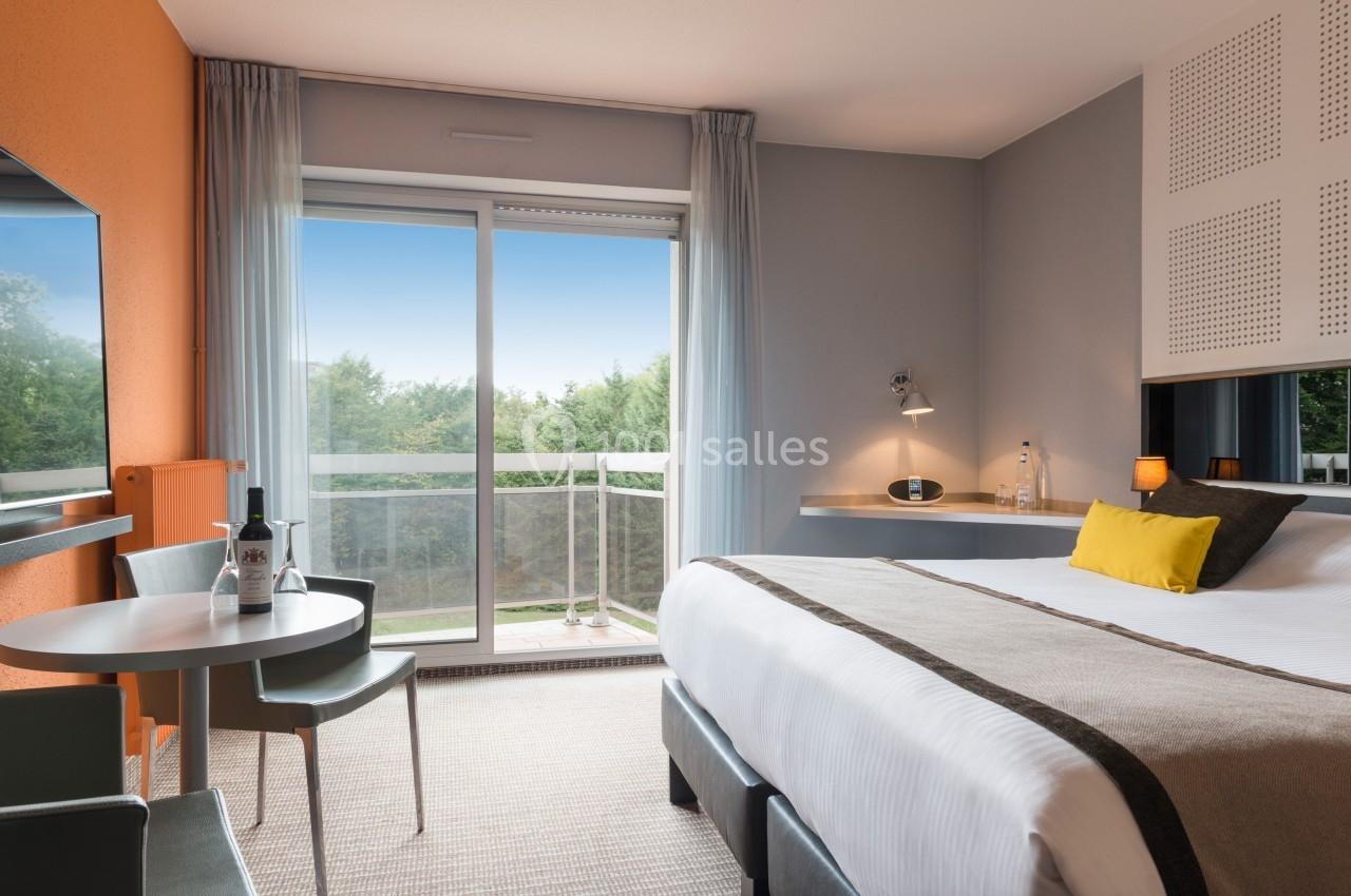 Chambre lumineuse avec lit double, table, chaises, balcon et vue sur des arbres à travers une baie vitrée.