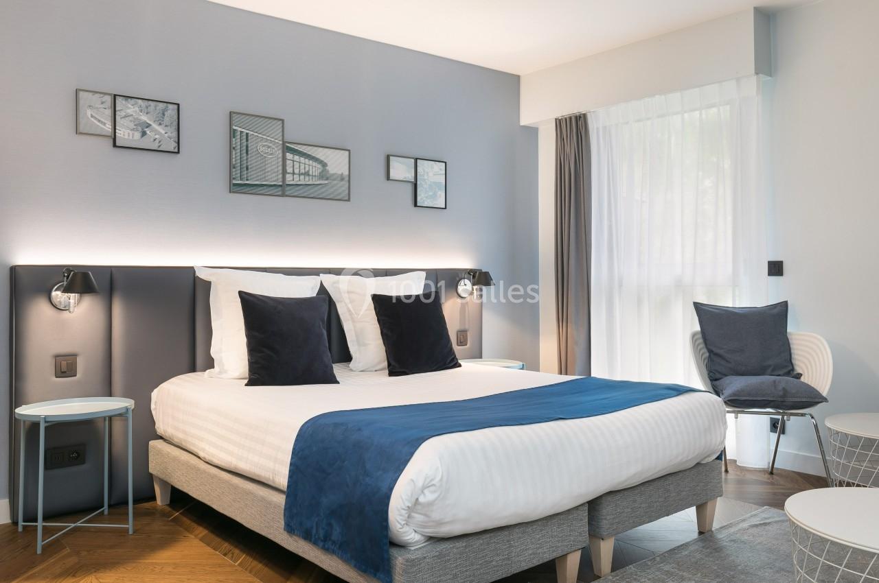 Chambre lumineuse avec un lit double, tête de lit rembourrée, coussins bleus, fauteuil et décor mural minimaliste.