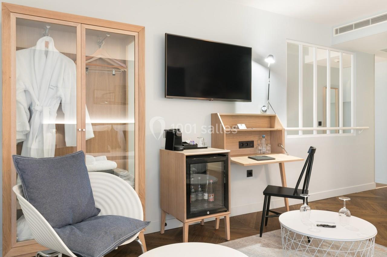 Chambre moderne avec coin salon, armoire vitrée, bureau avec minibar, télévision murale et éclairage intégré.