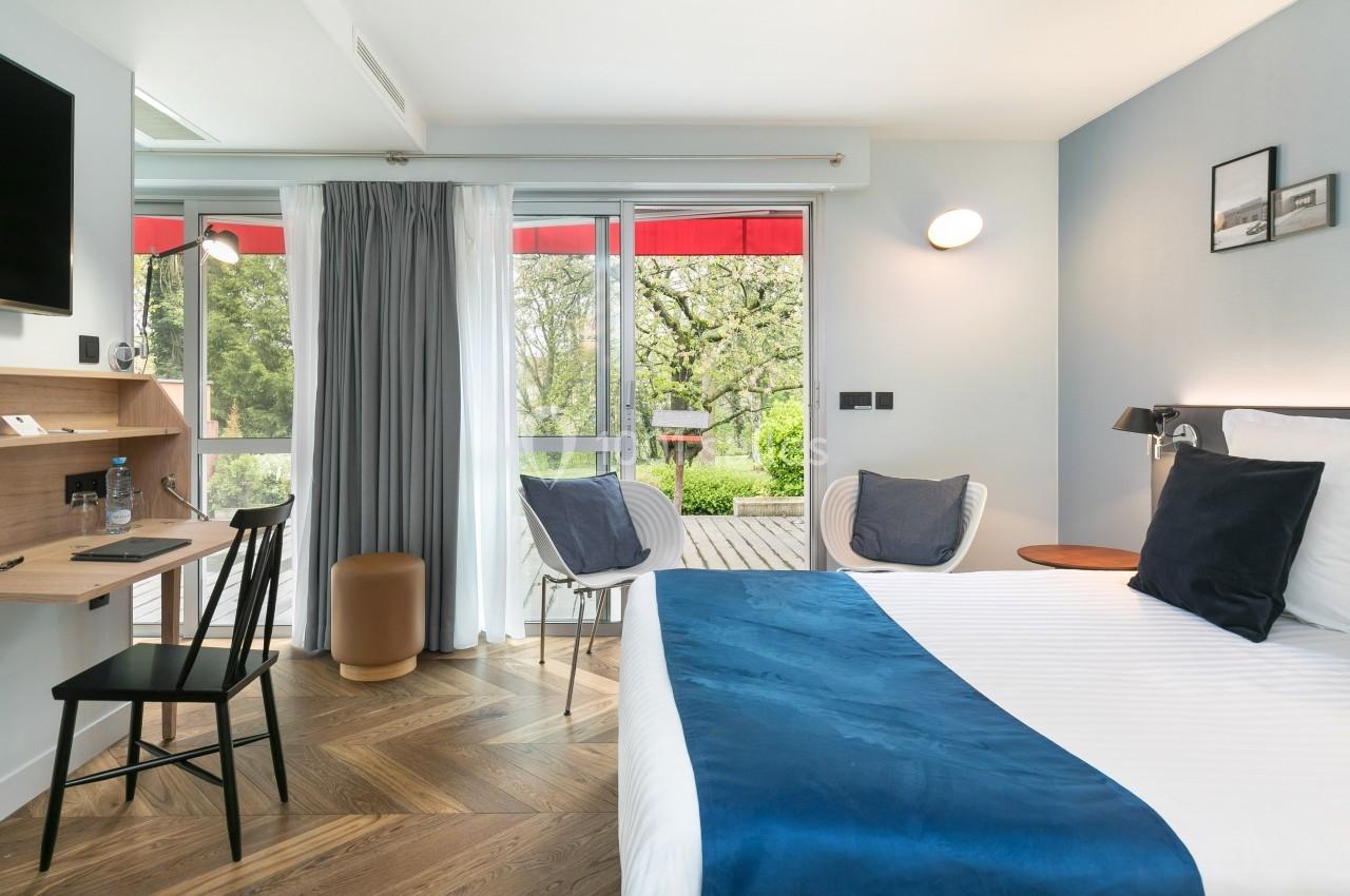 Chambre lumineuse avec lit double, bureau, fauteuils et grande baie vitrée donnant sur un balcon avec vue sur la verdure.