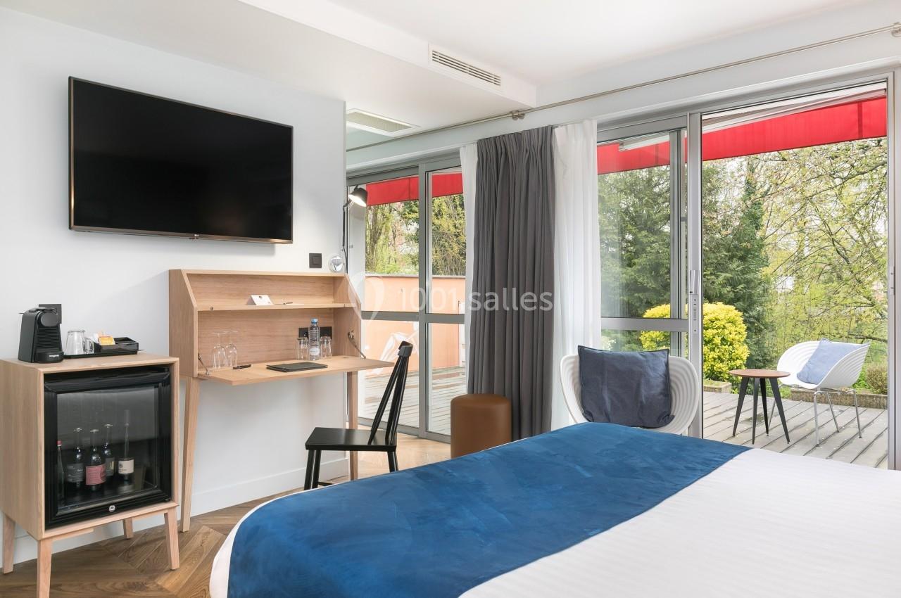 Chambre lumineuse avec lit, bureau, télévision, minibar et accès à une terrasse avec vue sur un jardin verdoyant.