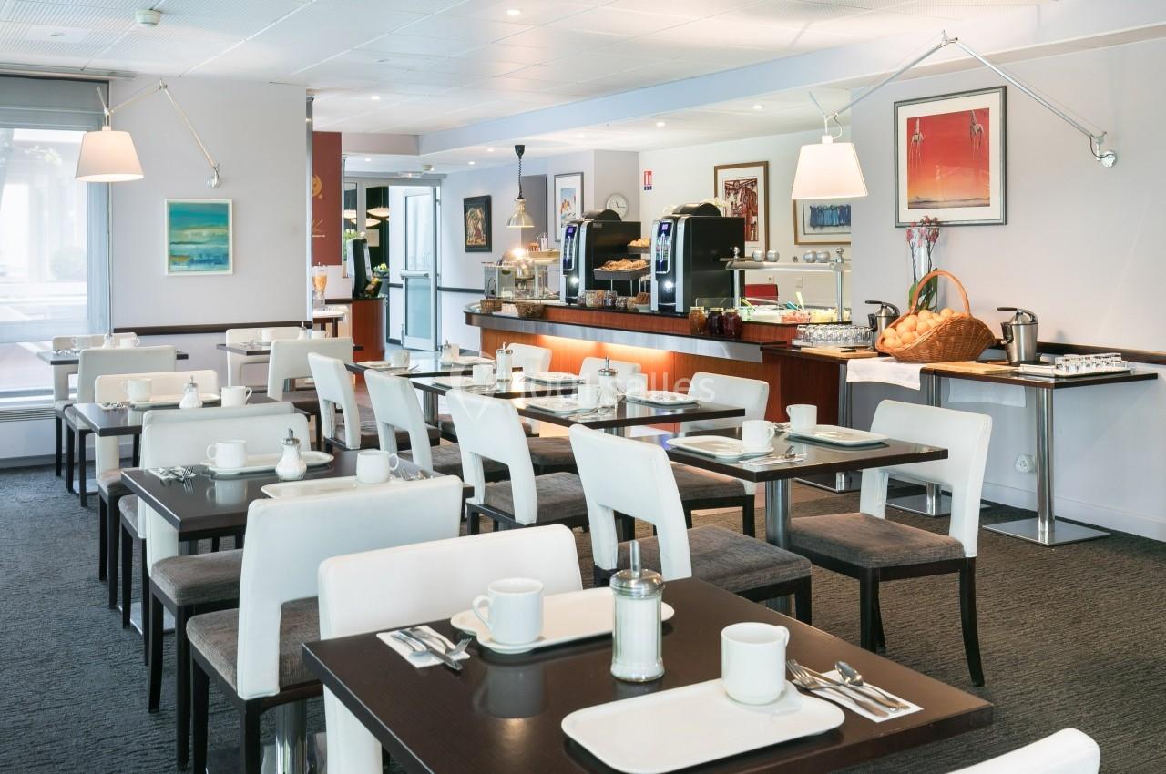 Salle de restaurant lumineuse avec tables dressées, chaises blanches et un buffet en arrière-plan.