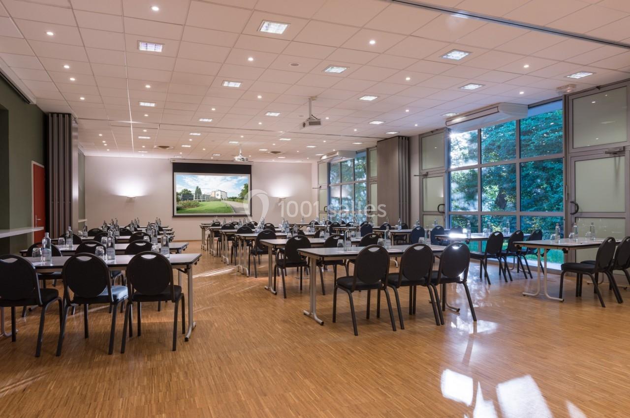 Salle de réunion lumineuse avec des tables et des chaises disposées, grandes fenêtres et écran mural.