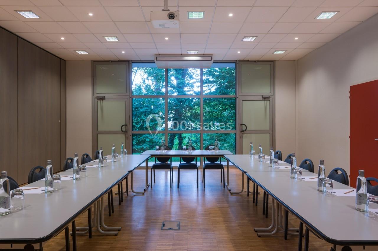 Salle de réunion lumineuse avec grandes fenêtres, tables disposées en U, chaises et bouteilles d'eau sur les tables.