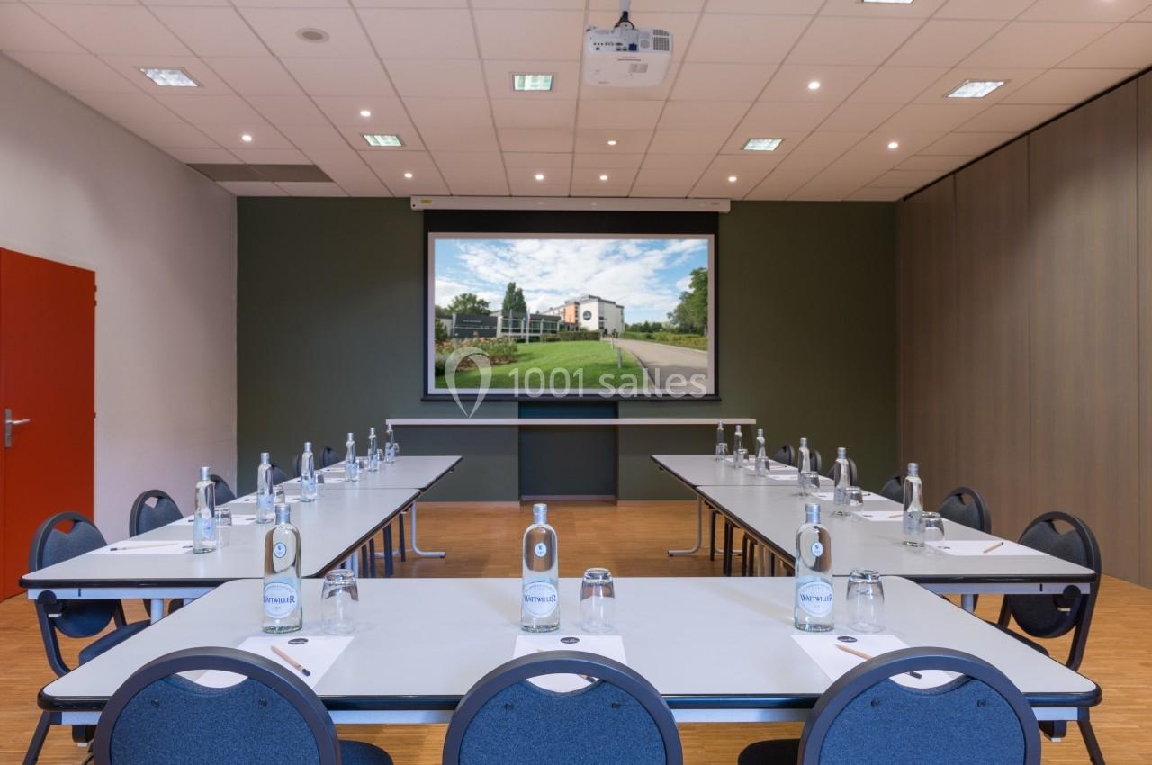 Salle de réunion équipée avec tables en U, chaises, écran de projection, bouteilles d'eau et éclairage au plafond.