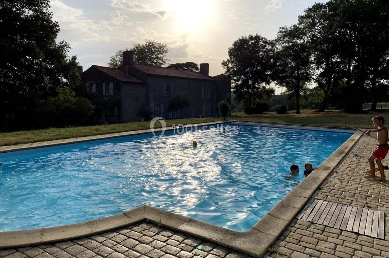 Piscine extérieure entourée d'arbres et d'une maison ancienne, avec des enfants jouant dans l'eau au coucher du soleil.