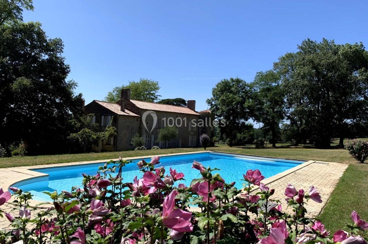 Piscine extérieure entourée de fleurs roses, avec une maison en pierre et des arbres en arrière-plan sous un ciel bleu.