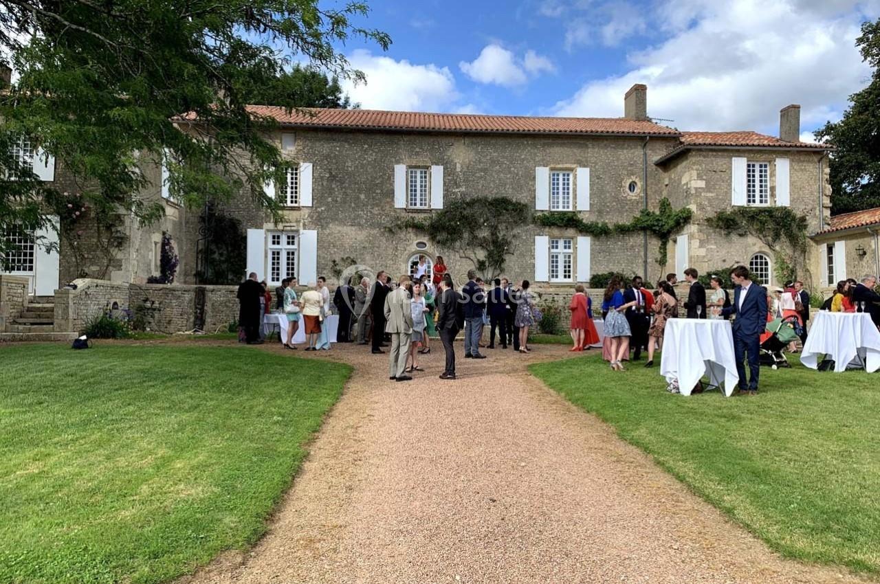 Groupe de personnes rassemblées devant une grande maison en pierre avec pelouse et ciel partiellement nuageux.