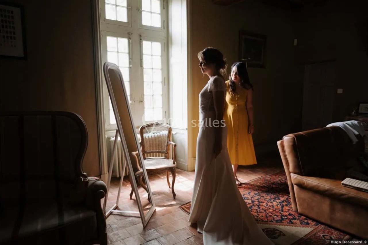 Une femme en robe de mariée se regarde dans un miroir, accompagnée d'une autre femme en robe jaune dans une pièce lumineuse.
