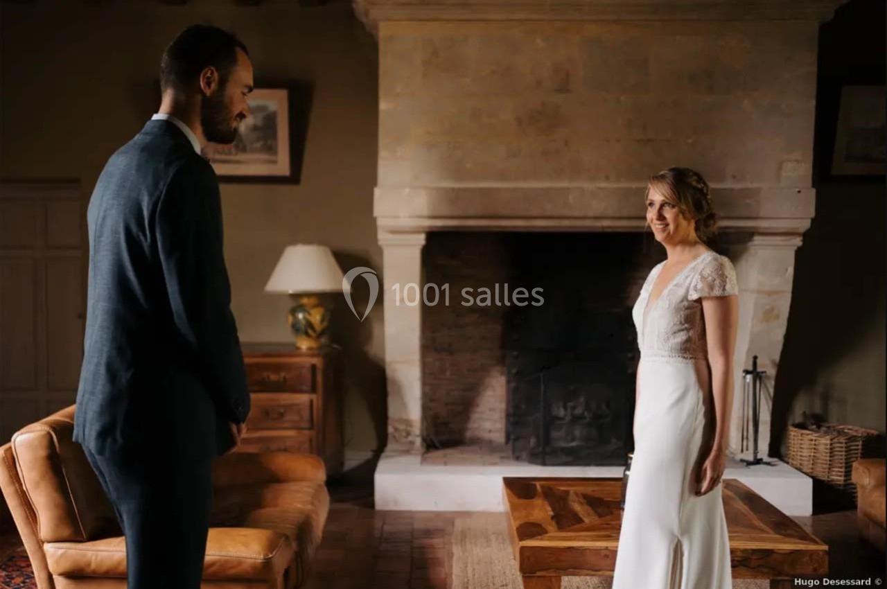 Un couple en tenue de mariage se regarde dans un salon avec une cheminée en pierre en arrière-plan.
