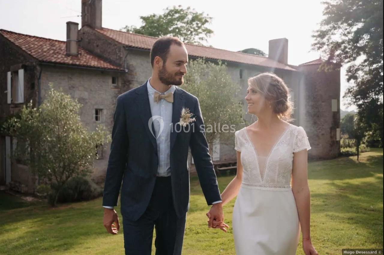 Un couple en tenue de mariage marche main dans la main dans un jardin, devant une maison en pierre.