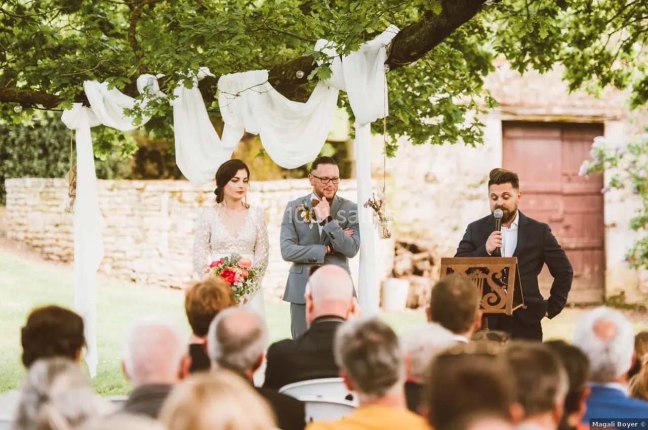 Un couple écoute un discours lors d'une cérémonie en plein air sous un arbre décoré de voilages blancs.