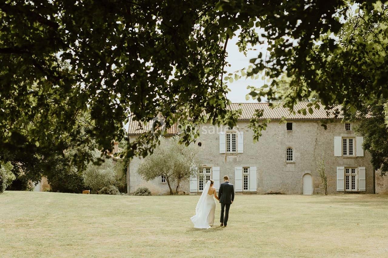 Un couple marche dans un jardin verdoyant devant une grande bâtisse en pierre sous un ciel dégagé.