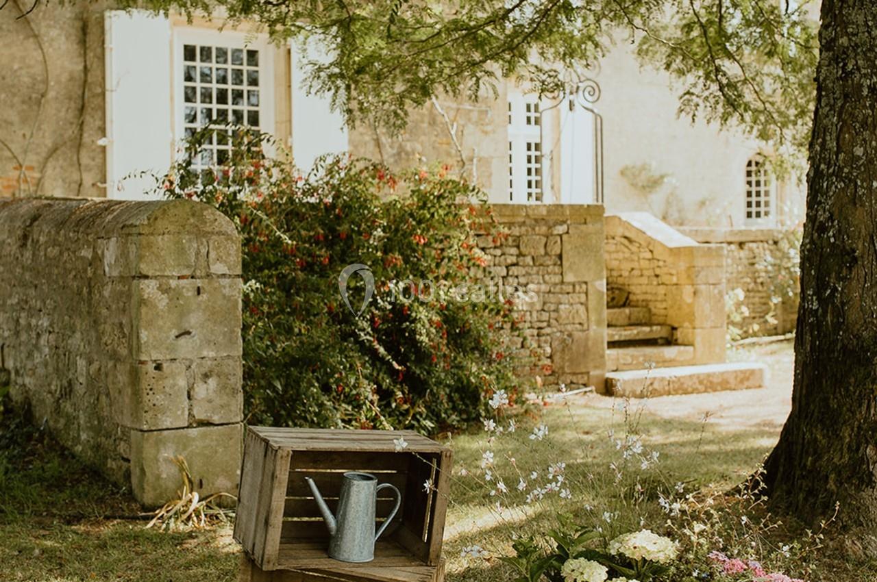 Arrosoir en métal posé sur une caisse en bois dans un jardin verdoyant devant une maison en pierre ancienne.