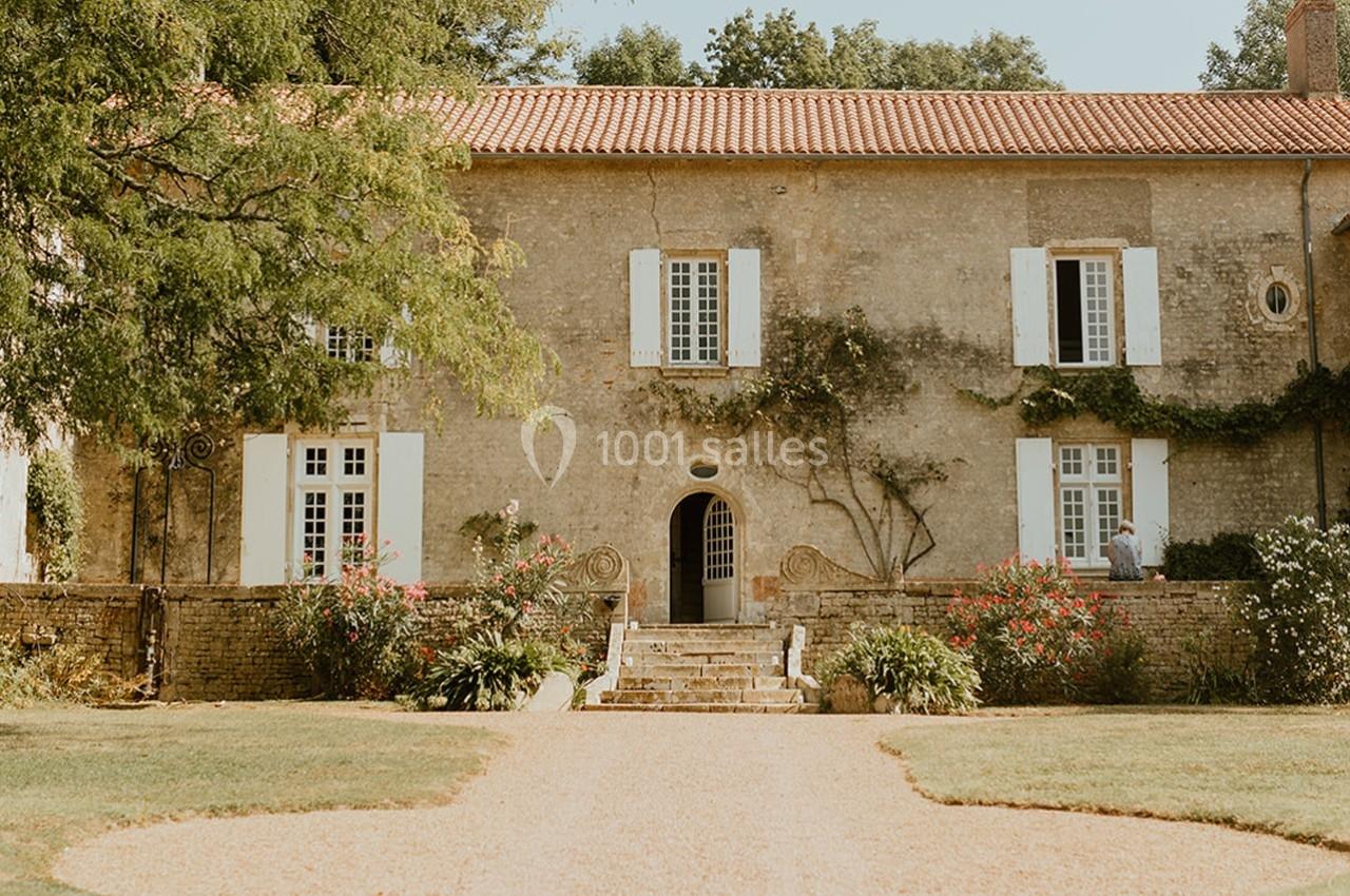Façade d'une maison en pierre avec volets blancs, entourée de végétation et d'un jardin avec allée gravillonnée.