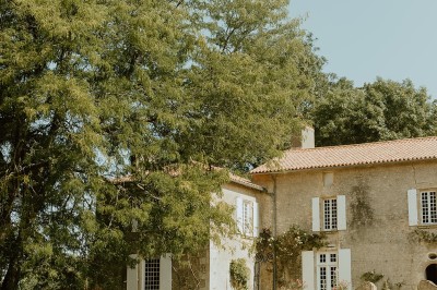 Miniature Location salle L'Orbrie (Vendée) - Le Logis des Arçonnières #68 Un couple marche dans un jardin verdoyant en direction d'une grande maison en pierre avec des volets blancs.