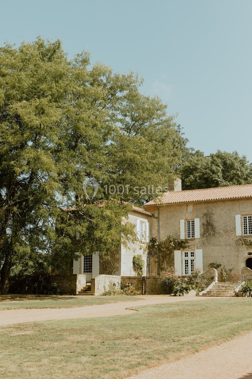 Grande maison en pierre avec volets blancs, entourée d'arbres et d'un jardin, sous un ciel dégagé.