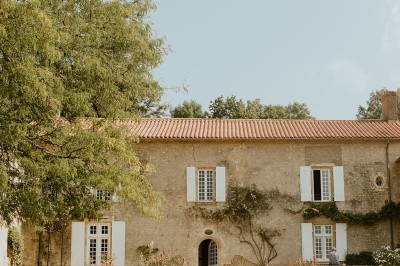 Miniature Location salle L'Orbrie (Vendée) - Le Logis des Arçonnières #69 Un couple marche dans un jardin verdoyant en direction d'une grande maison en pierre avec des volets blancs.