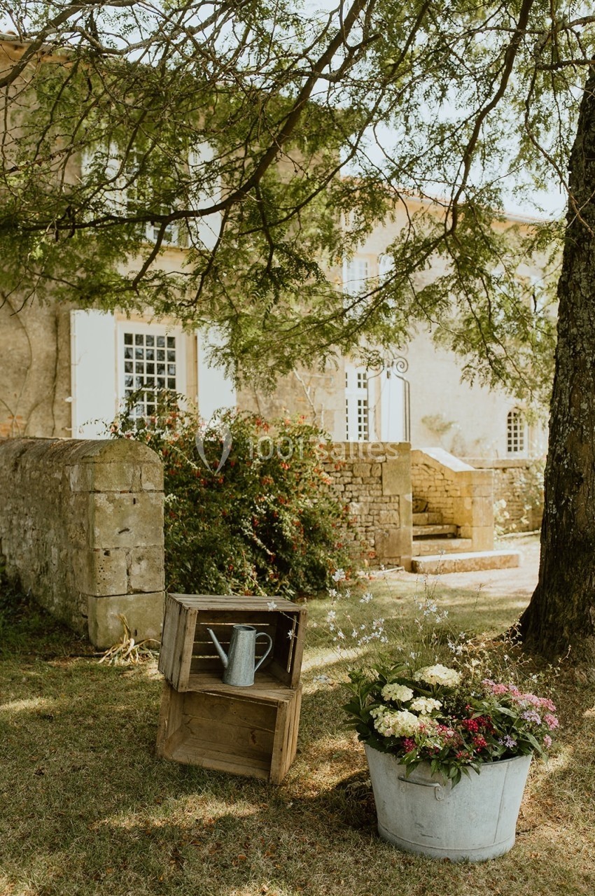 Jardin avec un seau de fleurs et une caisse en bois devant une maison en pierre aux volets blancs.