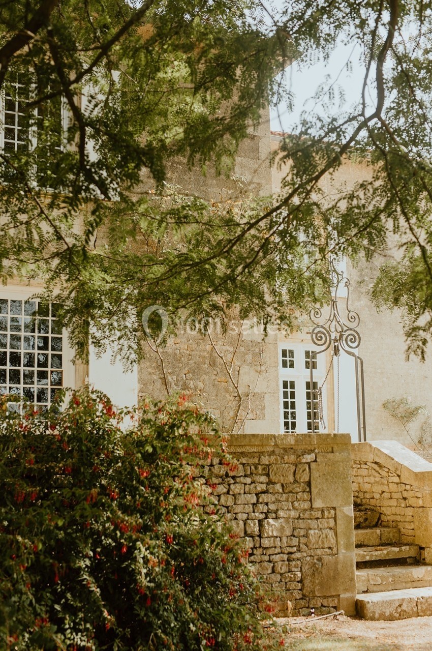 Façade en pierre d'un bâtiment ancien entouré de verdure, avec un portail en fer forgé et des arbustes fleuris.