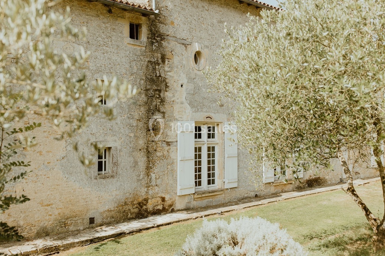Façade en pierre d'une maison ancienne avec des volets blancs, entourée d'arbres et d'un jardin verdoyant.