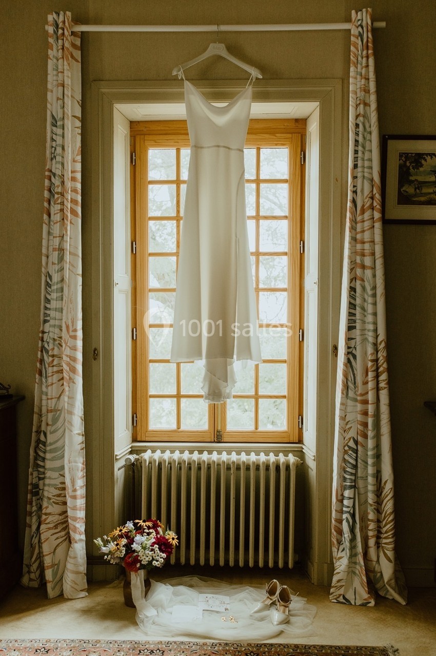 Robe de mariée blanche suspendue devant une fenêtre, avec bouquet, chaussures et accessoires posés au sol.