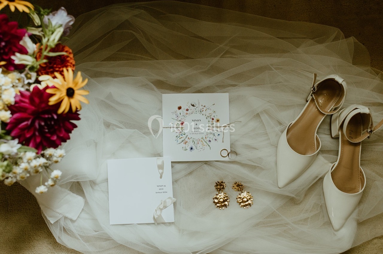 Carte d'invitation florale, chaussures blanches, bijoux dorés et bouquet coloré posés sur un voile de mariée.